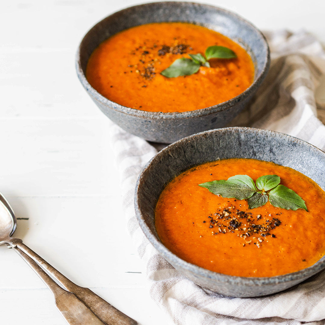 Two bowls of Tuscan Tomato Soup (Kit), topped with fresh basil and cracked black pepper, sit on a striped napkin with two spoons nearby, inviting you to enjoy the classic flavors of Tuscany.
